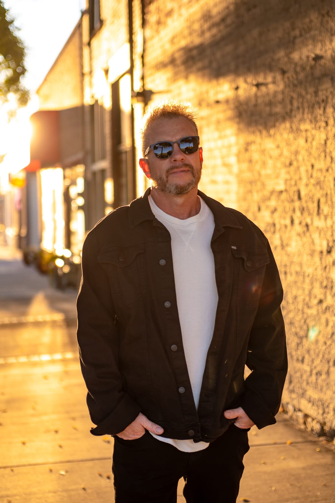Cecil Hawthorne at golden hour in a sunlit alley, in a dark jacket and sunglasses.